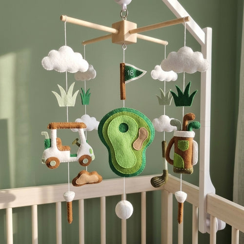 A baby nursery golf-themed mobile hanging in a white crib. The mobile features felt golf club, golf ball, flag, grass, and a green golf course target, suspended from a wooden crossbar with white cloud and grass shapes.
