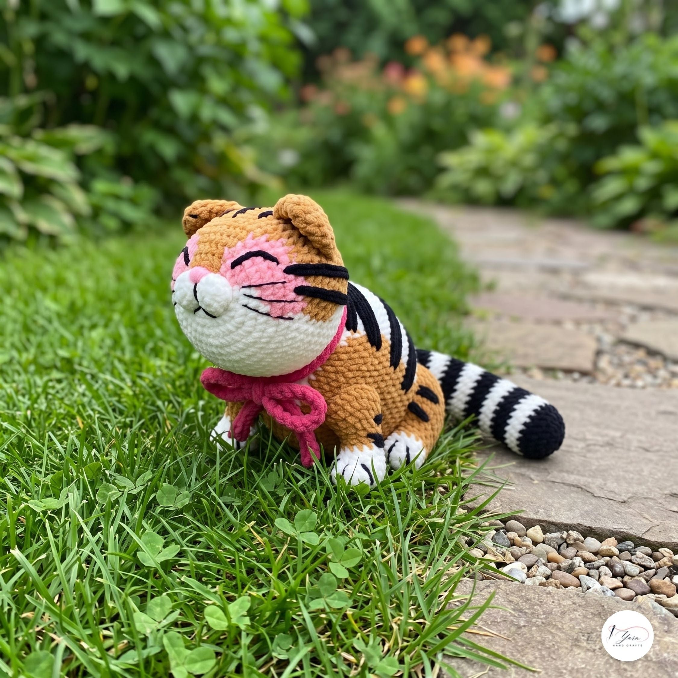 A crocheted tiger toy sitting on green grass beside a stone path in a garden setting.