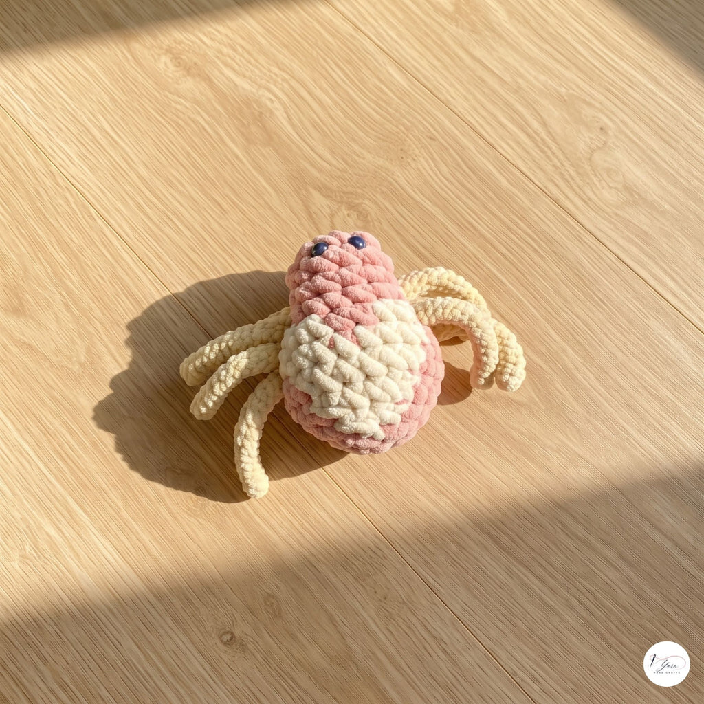 A close-up view of a plush handmade crochet spider toy with a pink and cream body, beige legs, and black safety eyes, sitting on a light wooden floor in warm natural sunlight.