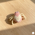A close-up view of a plush handmade crochet spider toy with a pink and cream body, beige legs, and black safety eyes, sitting on a light wooden floor in warm natural sunlight.