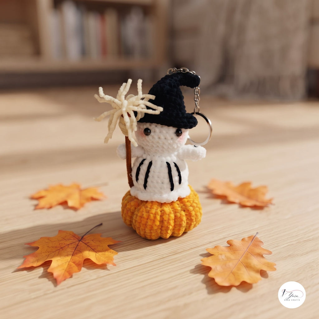 A crocheted witch keychain charm standing on a yellow pumpkin base, holding a broom, displayed on a wooden surface with scattered autumn leaves. The background is a softly blurred indoor setting.