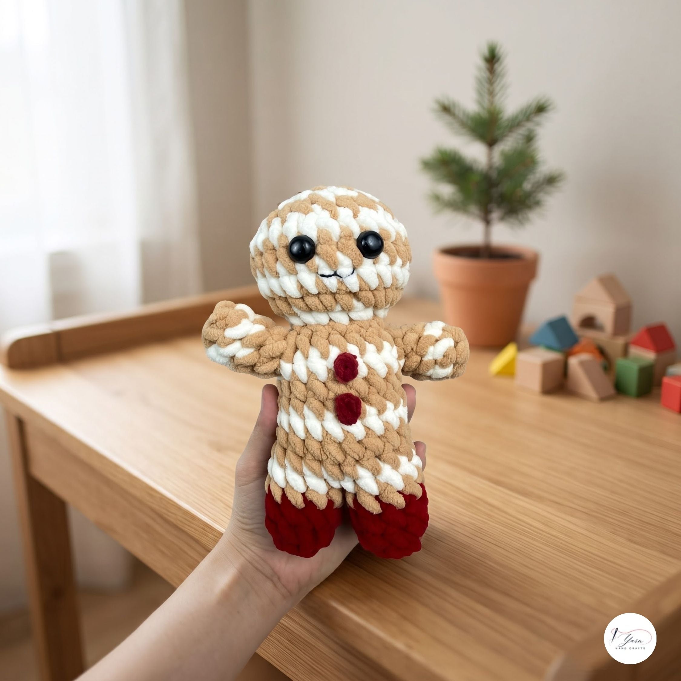 A handmade crocheted gingerbread person doll is held up by a person's hand over a light wood table in a softly lit room. The doll has beige and white striped yarn body, red yarn buttons and feet, and black yarn eyes.