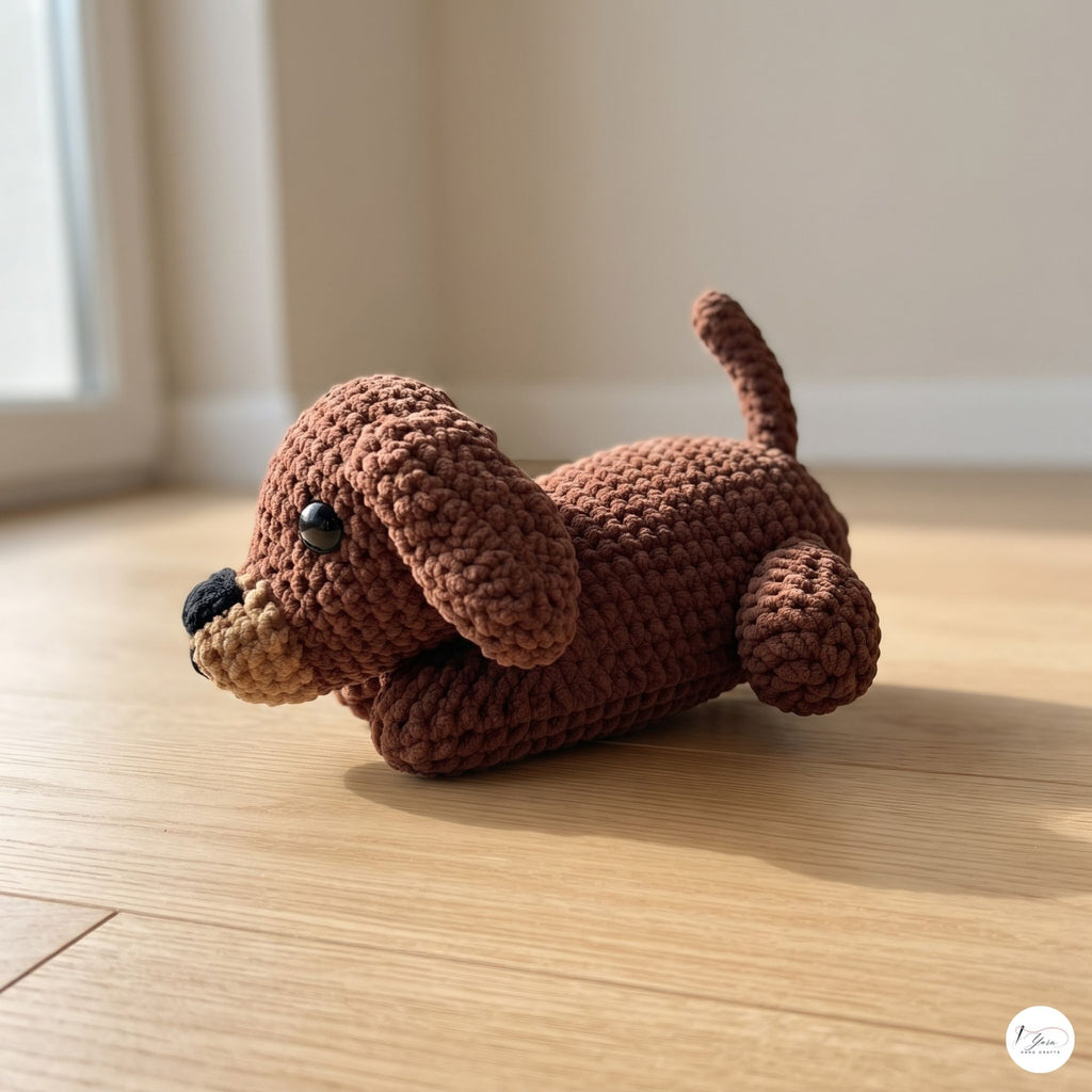 A small crocheted dog toy in medium brown with a beige snout, black nose, and black eyes, lying on a light wood floor in a softly lit room.