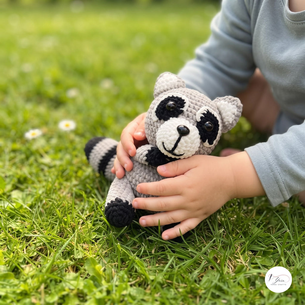 A child in a light blue long-sleeve shirt holds a crocheted raccoon toy on a green grass lawn. The raccoon is gray with black and white details.