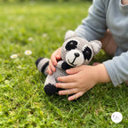 A child in a light blue long-sleeve shirt holds a crocheted raccoon toy on a green grass lawn. The raccoon is gray with black and white details.