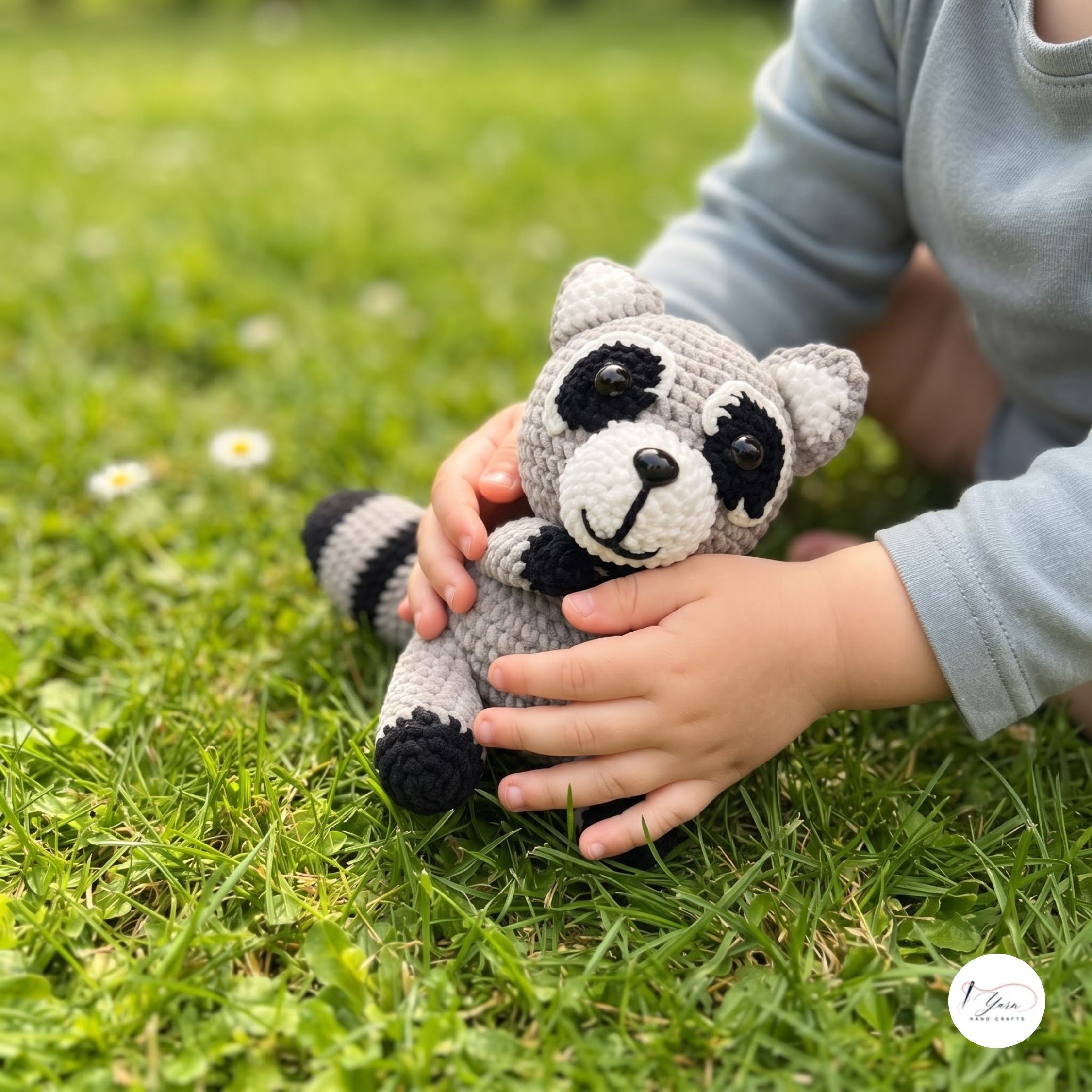 A child in a light blue long-sleeve shirt holds a crocheted raccoon toy on a green grass lawn. The raccoon is gray with black and white details.