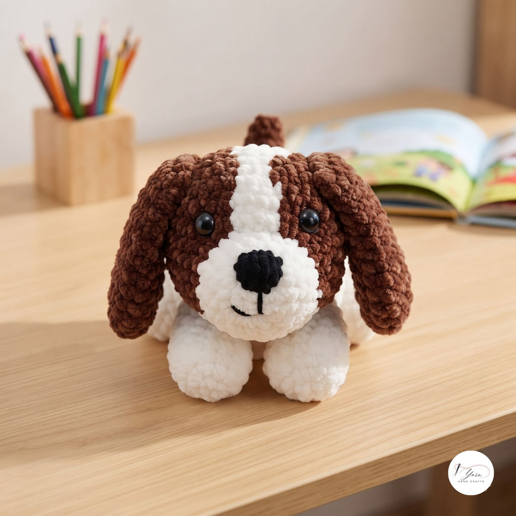A crocheted dog toy with brown and white yarn sits on a light wooden desk, with a pencil holder and an open book blurred in the background.