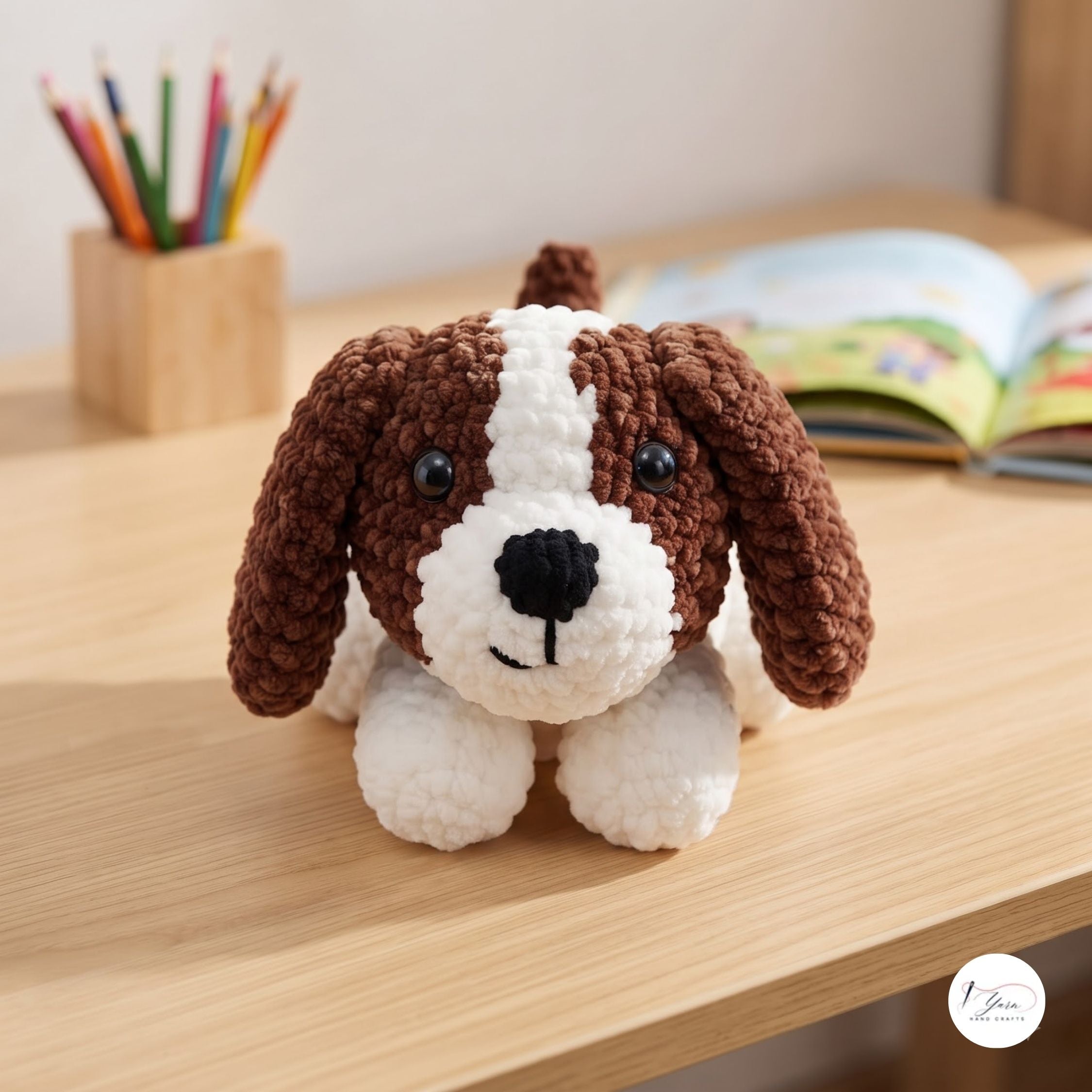 A crocheted dog toy with brown and white yarn sits on a light wooden desk, with a pencil holder and an open book blurred in the background.