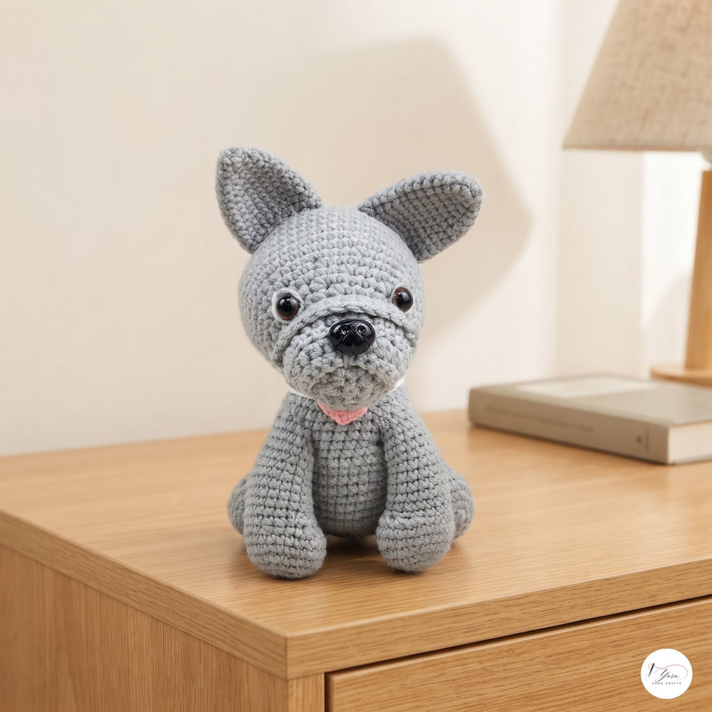 A crocheted gray dog toy with black eyes and nose and a small pink collar, sitting on a light wood table in a softly lit room.