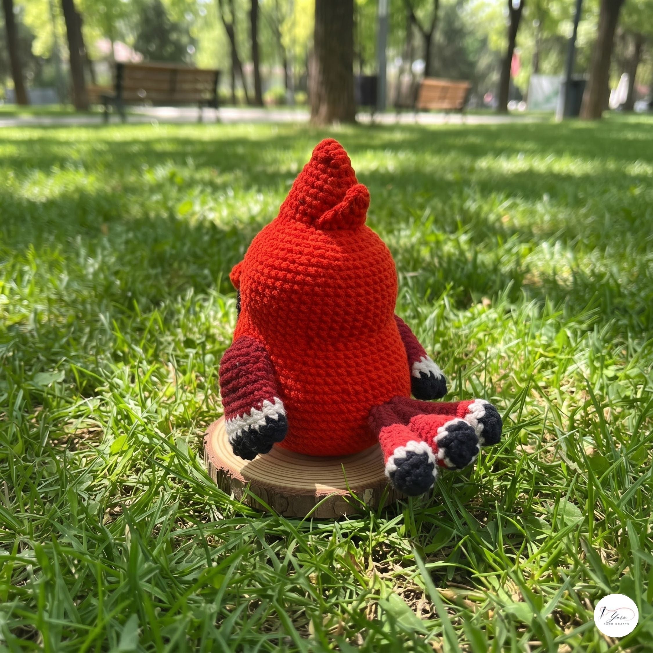 A red crocheted plush toy with black and white striped feet sits on a small round wooden slice in a grassy park setting.