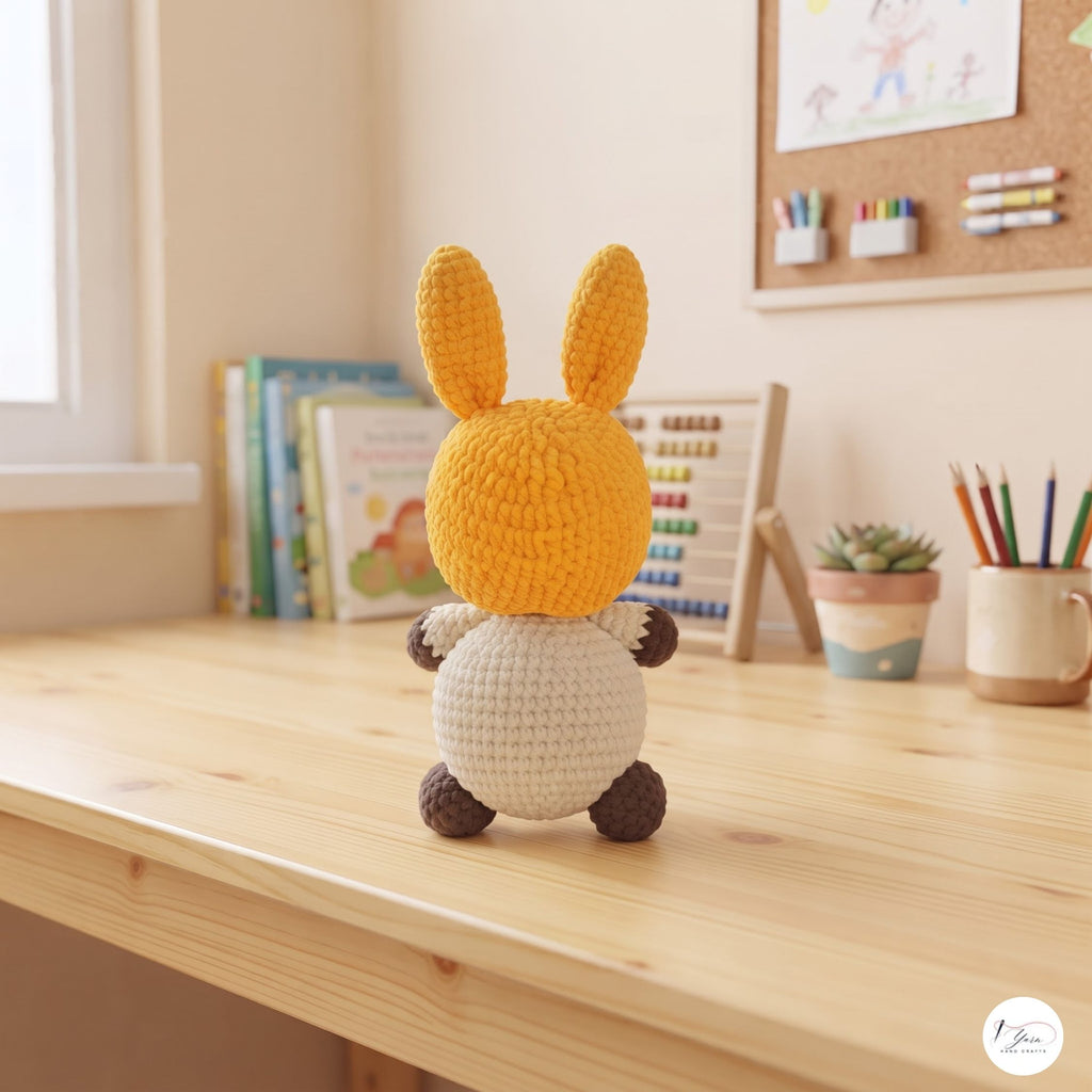 A crocheted bunny plush toy with a yellow-orange head and ears, a cream body, and dark brown paws and feet, sitting on a light wooden desk in a bright, cozy room.