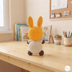 A crocheted bunny plush toy with a yellow-orange head and ears, a cream body, and dark brown paws and feet, sitting on a light wooden desk in a bright, cozy room.