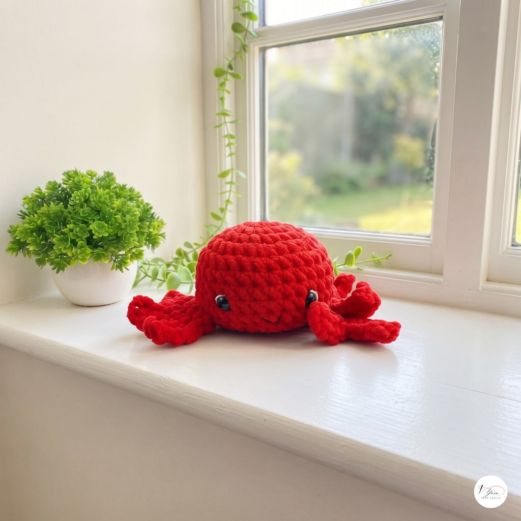 A red crocheted octopus plush toy with black bead eyes, resting on a white windowsill next to a small potted green plant.