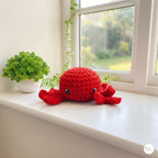 A red crocheted octopus plush toy with black bead eyes, resting on a white windowsill next to a small potted green plant.