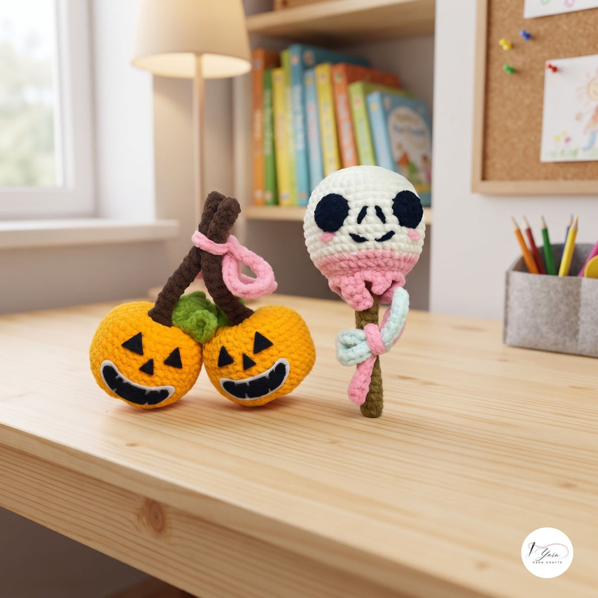 Three handmade crocheted Halloween-themed ornaments displayed on a light wood desk in a cozy room setting. The set includes two orange jack-o'-lanterns on a brown stem with pink loops, and a white skull on a pink and mint green staff.