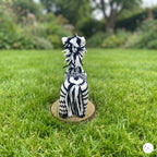 A handmade black and white knit zebra doll sitting on a round wooden base, photographed outdoors on a green grass lawn with a blurred garden background.