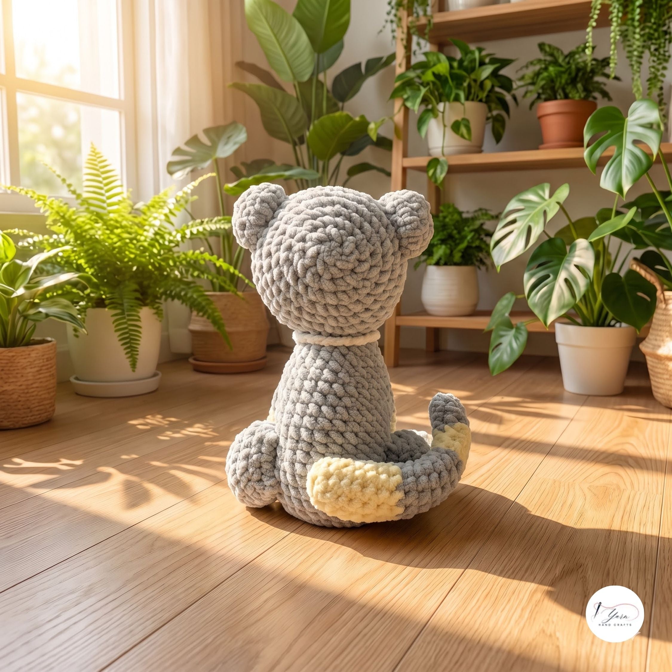 A crocheted plush bear toy sits on a wooden floor in a sunlit room filled with potted plants and shelves. The bear is light gray with cream-colored paws and tail tip, shown from the back.