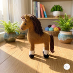 A crocheted toy horse with a brown mane and tail, tan body, and black-and-white hooves, standing on a wooden floor in a sunlit room with potted ferns and books in the background.