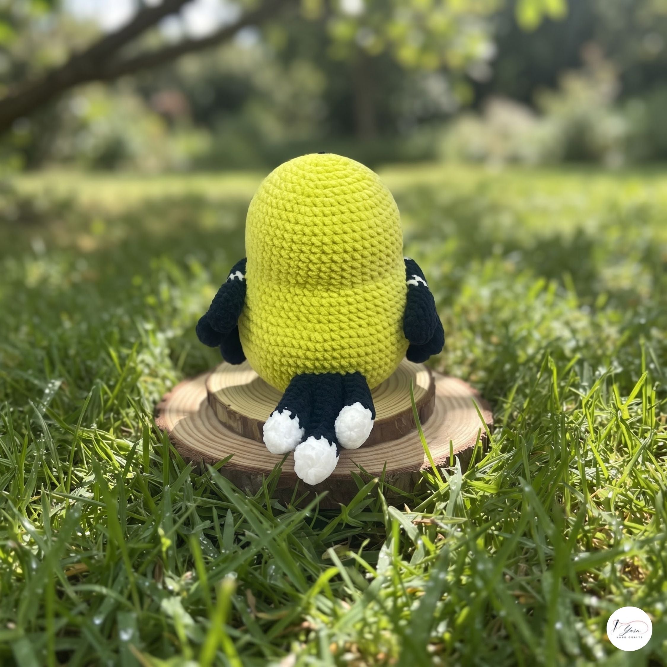 A crocheted plush toy shaped like a penguin sitting on a round wooden slice in a grassy outdoor setting. The plush has a bright yellow body, black wings and legs, and white feet.