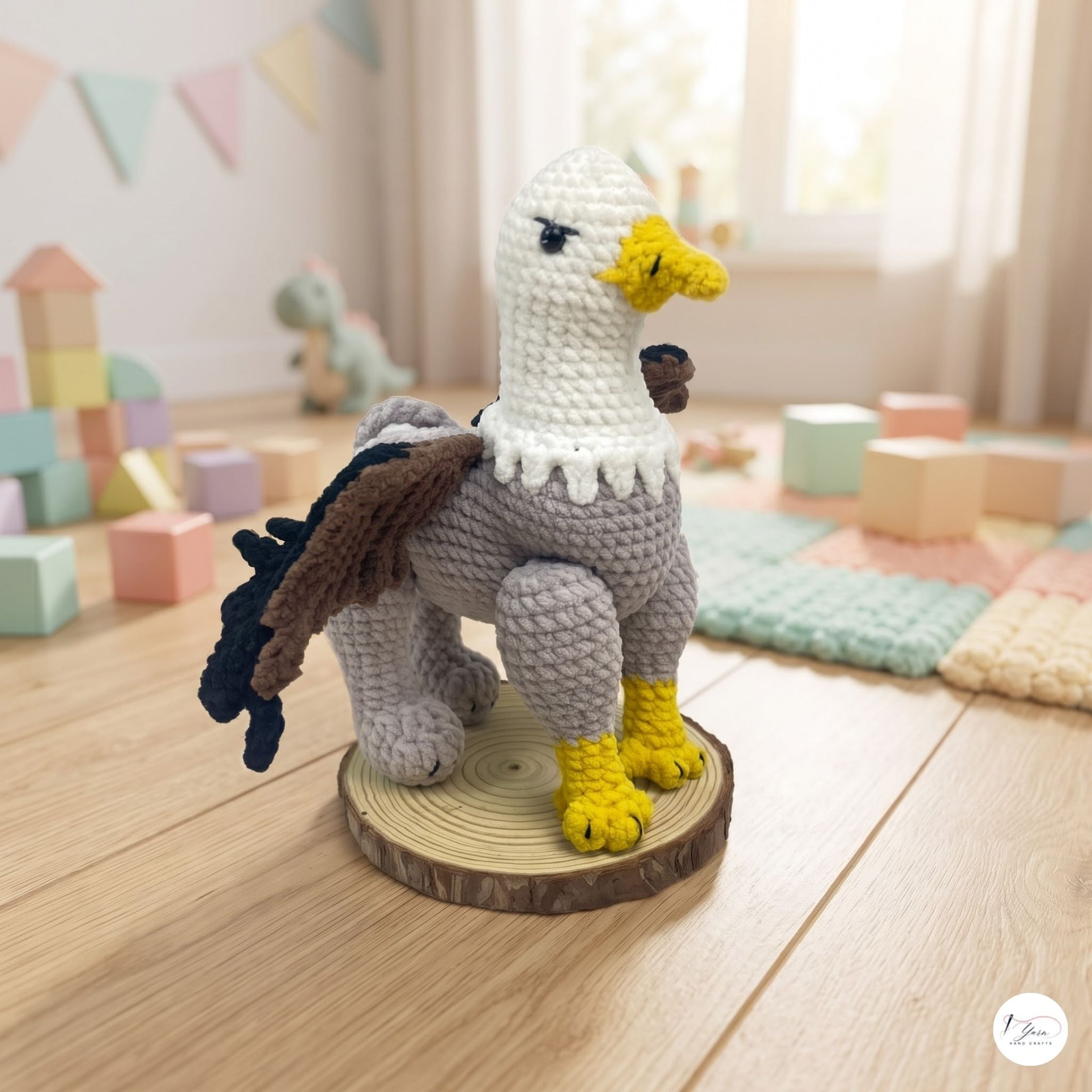 A crocheted eagle figurine standing on a round wood slice base, photographed in a softly lit children's room with pastel blocks and toys in the background.