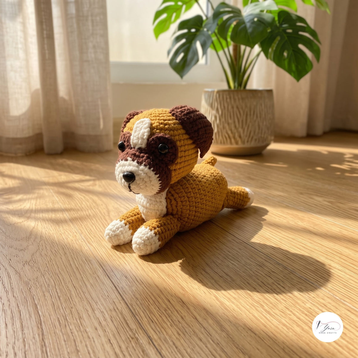 A crocheted plush puppy toy in brown, cream, and white, lying on a wooden floor in warm sunlight near a potted plant.
