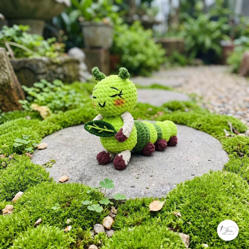 A handmade green crochet caterpillar toy holding a leaf, sitting on a round stone in a mossy garden setting.