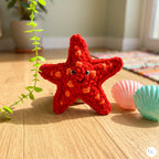 A red crocheted starfish toy with orange spots and a smiling face sits on a light wood floor, styled with pink and teal seashells and a green plant in a bright room.