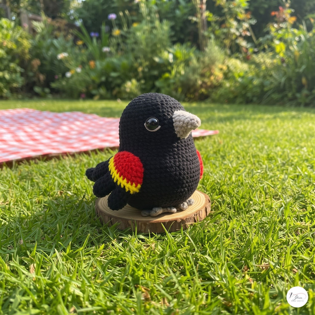 A crocheted black bird figurine with red and yellow wing accents and a gray beak, standing on a small wooden slice in a grassy outdoor garden setting.