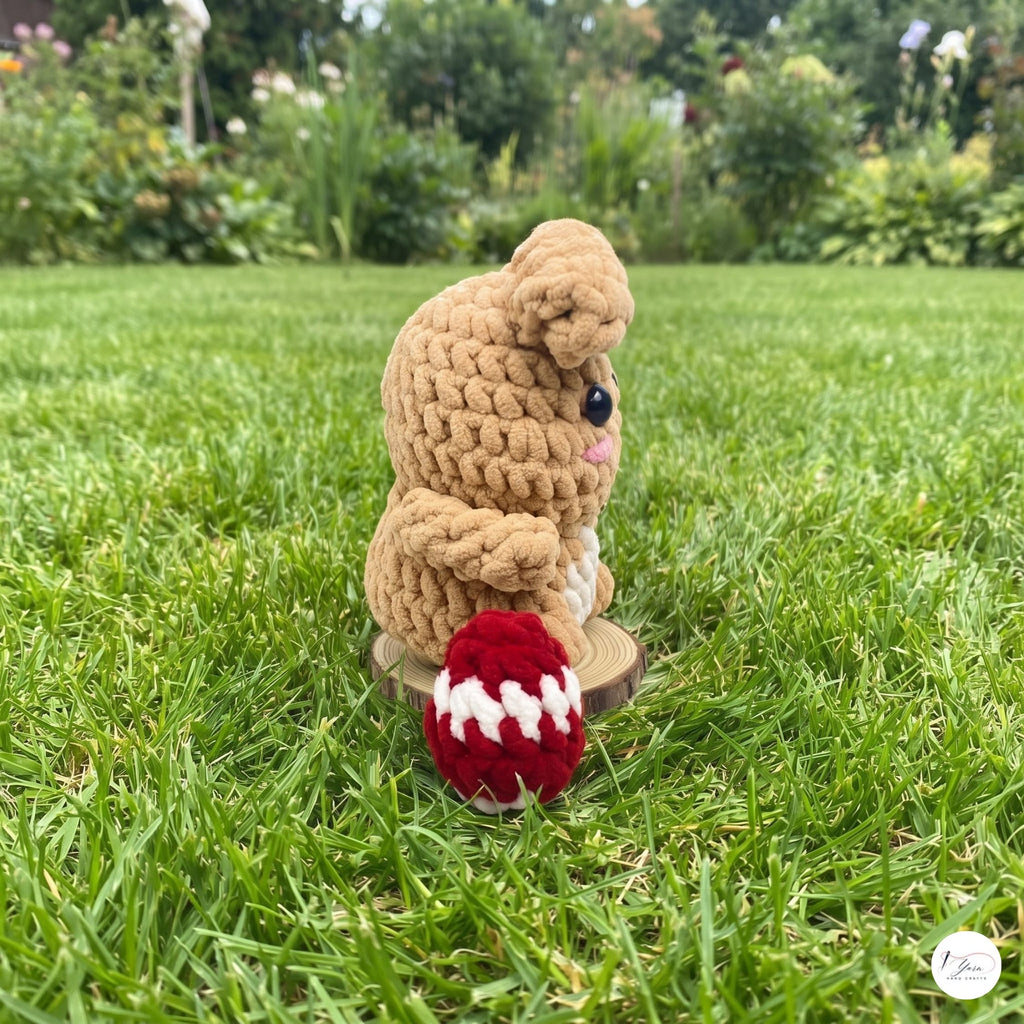 A small crocheted teddy bear figurine sits on green grass in a garden, holding a red and white crocheted egg. The background shows a blurred green lawn and foliage.
