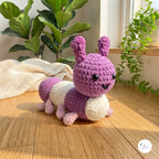 A crocheted caterpillar toy in purple and cream, posed on a wooden floor in a bright room with potted plants and a textured blanket.