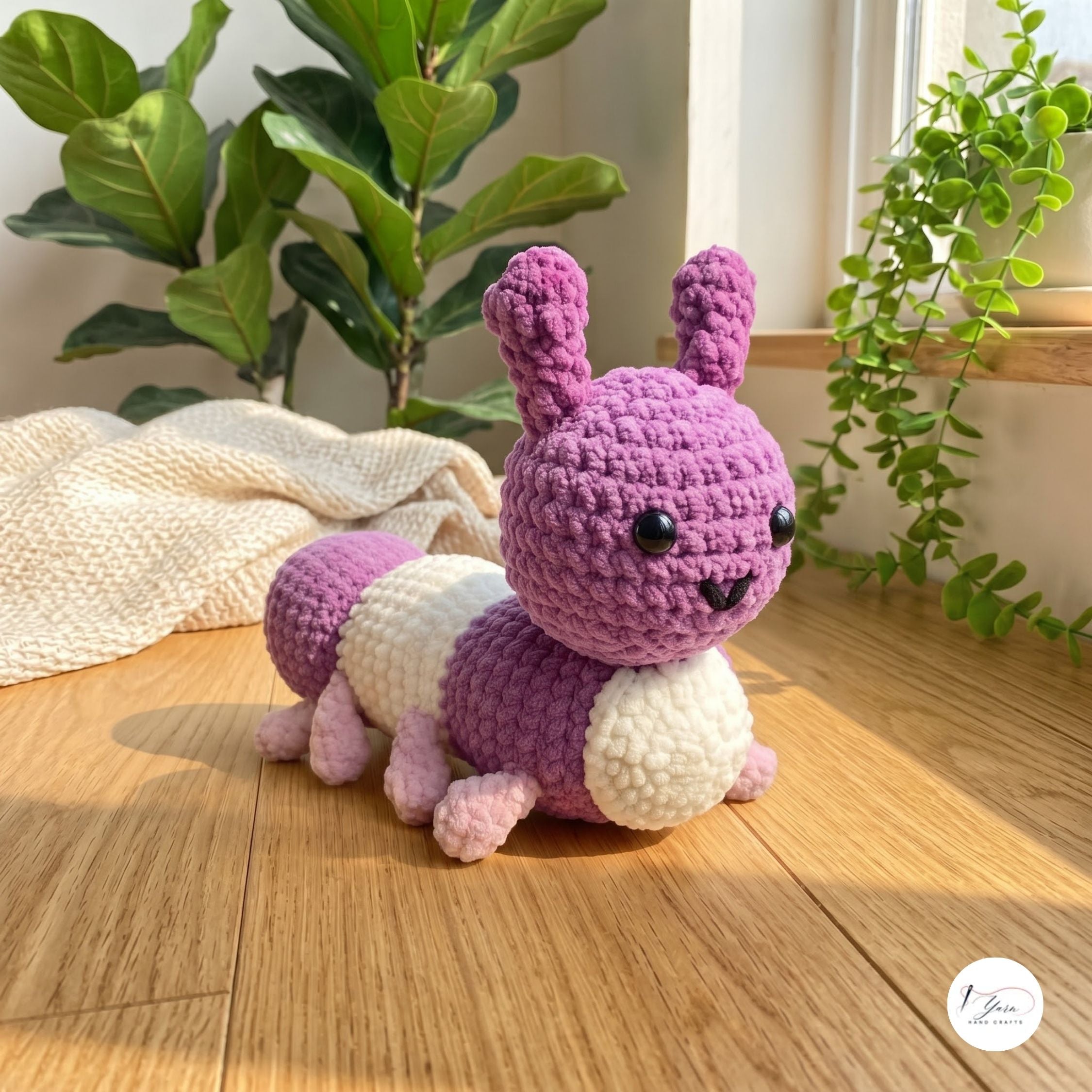 A crocheted caterpillar toy in purple and cream, posed on a wooden floor in a bright room with potted plants and a textured blanket.