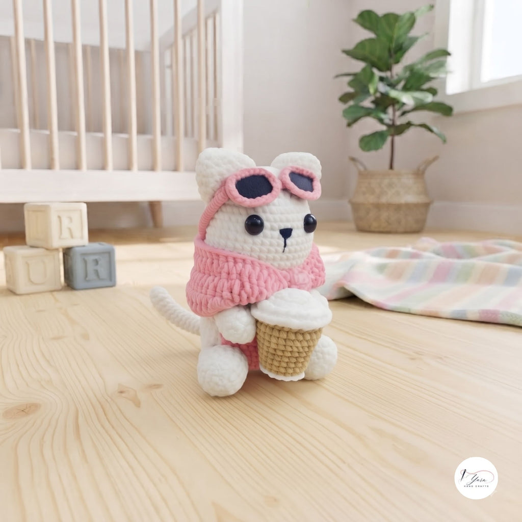 A white and pink crocheted plush cat toy wearing pink sunglasses and holding a cupcake, set on a light wood floor in a softly styled nursery with a crib, plant, and pastel blanket.