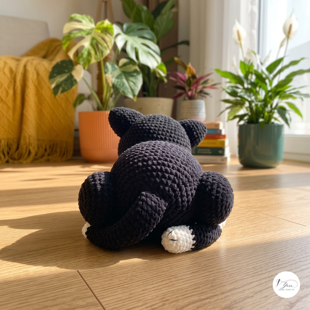 A black crocheted cat plush toy is shown from behind, sitting on a wooden floor in a brightly lit room with potted plants and a yellow blanket in the background.