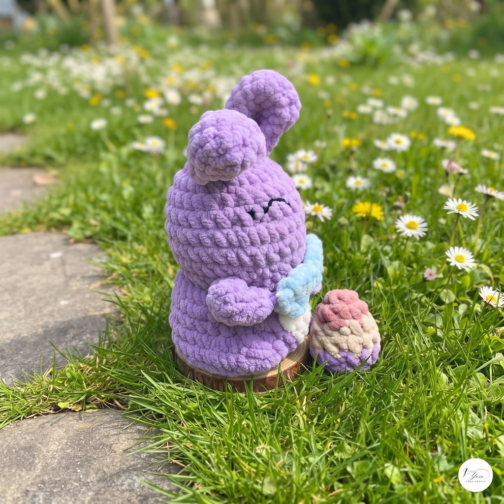 A purple crocheted bunny figure sitting on a grassy lawn with white and yellow flowers, holding a small pastel egg and a blue flower.