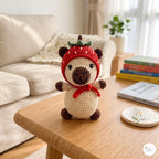 A crocheted bear doll wearing a red strawberry hat and red scarf, sitting on a wooden table in a cozy living room setting.