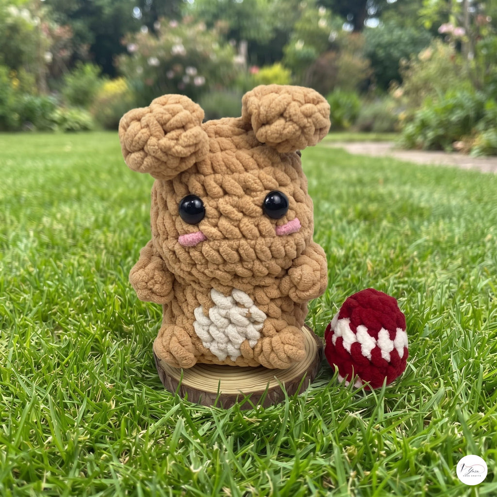 A crocheted plush bunny toy made from chunky tan yarn stands on a small wooden slice in a grassy garden, with a small red-and-white crocheted egg beside it.