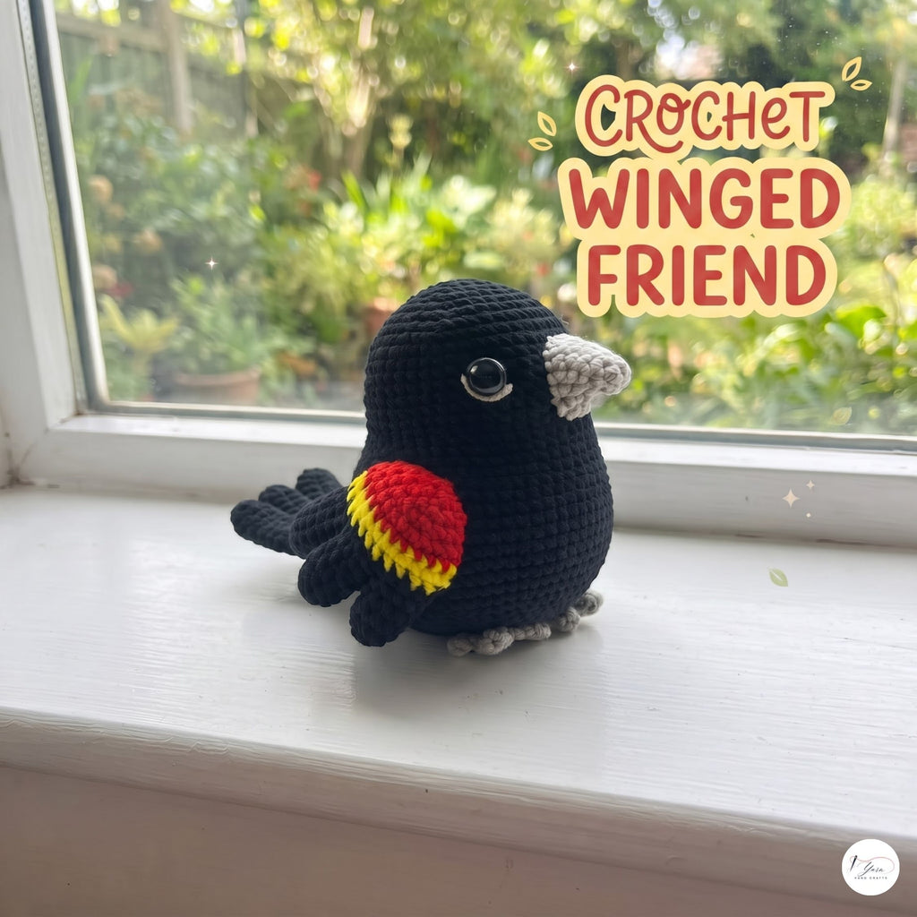 A crocheted bird toy with a black body, red and yellow wing detail, and gray beak and feet, sitting on a white windowsill with a garden view outside. Text in the upper right reads 'CROCHET WINGED FRIEND'.