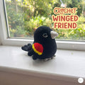 A crocheted bird toy with a black body, red and yellow wing detail, and gray beak and feet, sitting on a white windowsill with a garden view outside. Text in the upper right reads 'CROCHET WINGED FRIEND'.