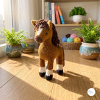 A crocheted horse toy standing on a wooden floor in a sunlit room, with potted ferns, books, and a basket of yarn in the background.