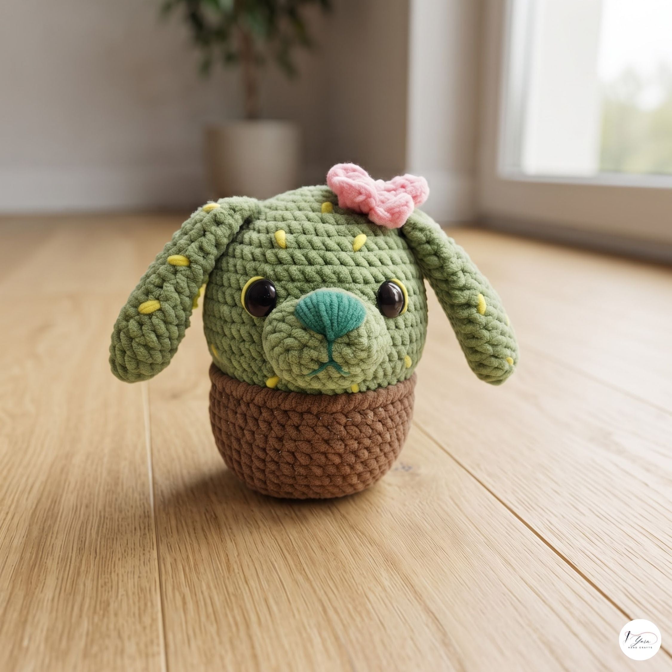 A front-facing plush handmade crochet cactus puppy toy with long floppy ears, yellow embroidered spines, a pink bow flower, and a turquoise nose, sitting in a brown crochet plant pot base on a sunlit wooden floor.