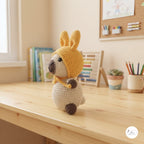A crocheted llama or alpaca doll sits on a light wooden desk in a bright, cozy room. The doll has a yellow crocheted hood with long ears, a cream-colored body, and dark brown accents on its snout, paws, and hooves.