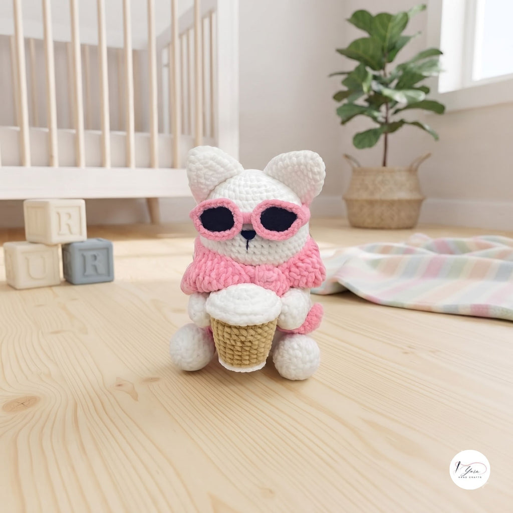 A cute handmade white crochet cat plush sitting on a light wooden floor in a cozy nursery, wearing a pink sweater and pink sunglasses over its eyes while holding a crocheted coffee cup.