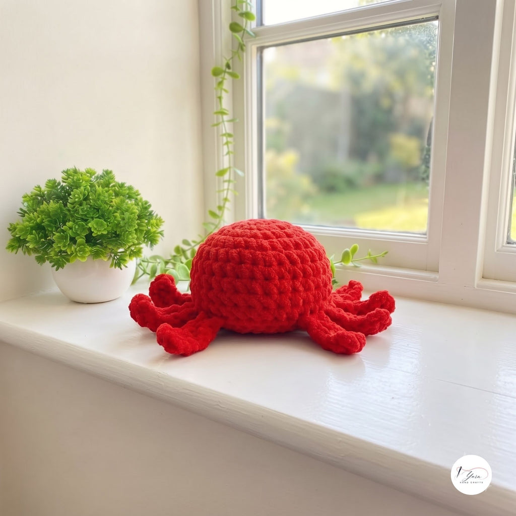 A bright red crocheted octopus plush toy sits on a white windowsill next to a small potted green plant, with a soft-focus garden visible outside the window.