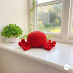 A bright red crocheted octopus plush toy sits on a white windowsill next to a small potted green plant, with a soft-focus garden visible outside the window.