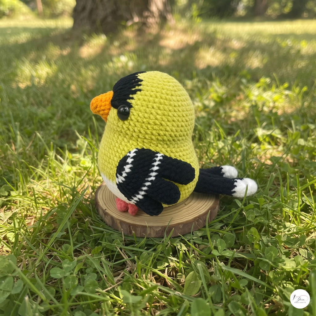 A crocheted bird toy in bright yellow-green with black, white, and orange details, standing on a small round wooden slice in a grassy outdoor setting.