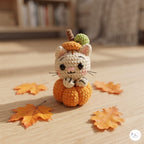 A crocheted amigurumi cat dressed as a pumpkin sits on a wooden surface with scattered autumn leaves. The toy has an off-white body, an orange pumpkin base, a small orange hat with a brown stem, and a green leaf on top.
