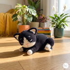 A crocheted black and white cat toy lying on a wooden floor in a sunlit room with potted plants and a yellow blanket in the background.