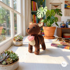 A crocheted brown cat figurine holding a pink heart-shaped accessory, displayed on a sunlit windowsill in a cozy room with potted plants, books, and decor.