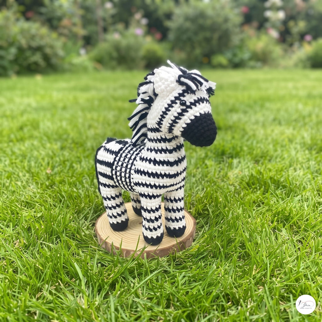 A crocheted zebra toy standing on a round wooden base in a grassy garden setting.