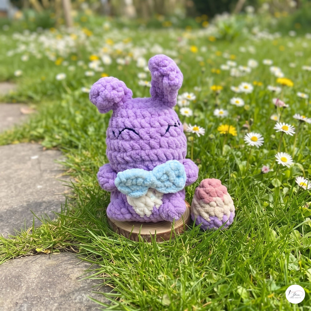 A crocheted purple bunny figurine with long ears, embroidered closed eyes, and a light blue bow tie, standing on a small wooden base in a grassy garden with white and yellow flowers. A matching small crocheted egg sits beside it.